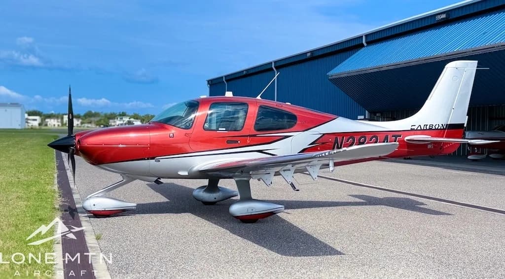 A red and silver Cirrus SR22T aircraft, registration N282AT, parked on an tarmac next to a blue hangar.