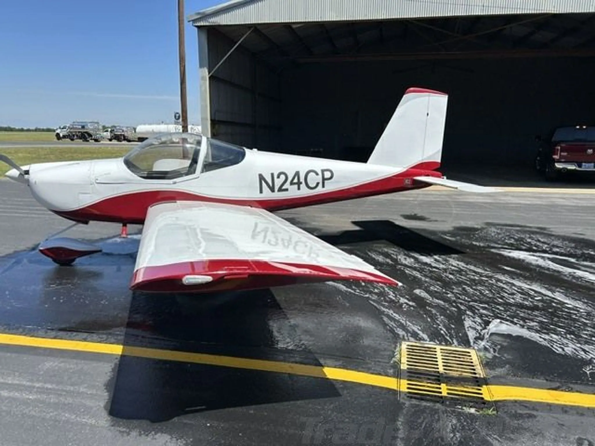 A white and red Van's RV-12 aircraft, N24CP, is parked on a wet tarmac outside a hangar.