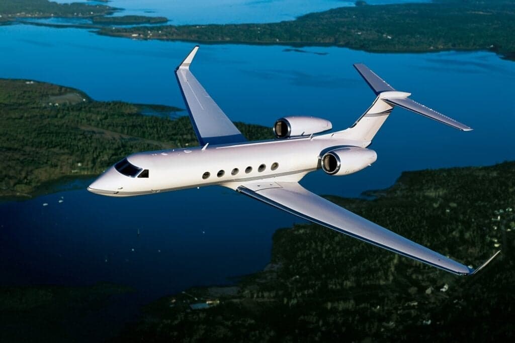Gulfstream business jet in flight over a forested landscape and blue water.