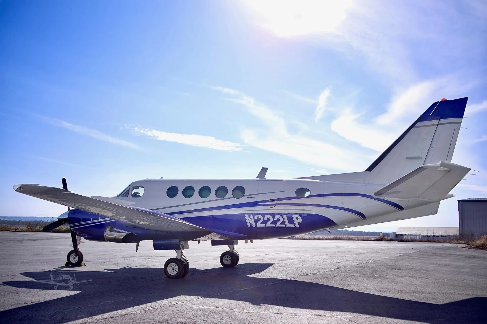 A white and blue twin-engine turboprop aircraft (Beechcraft King Air) with registration N222LP parked on an asphalt ramp, with the sun shining brightly from behind.