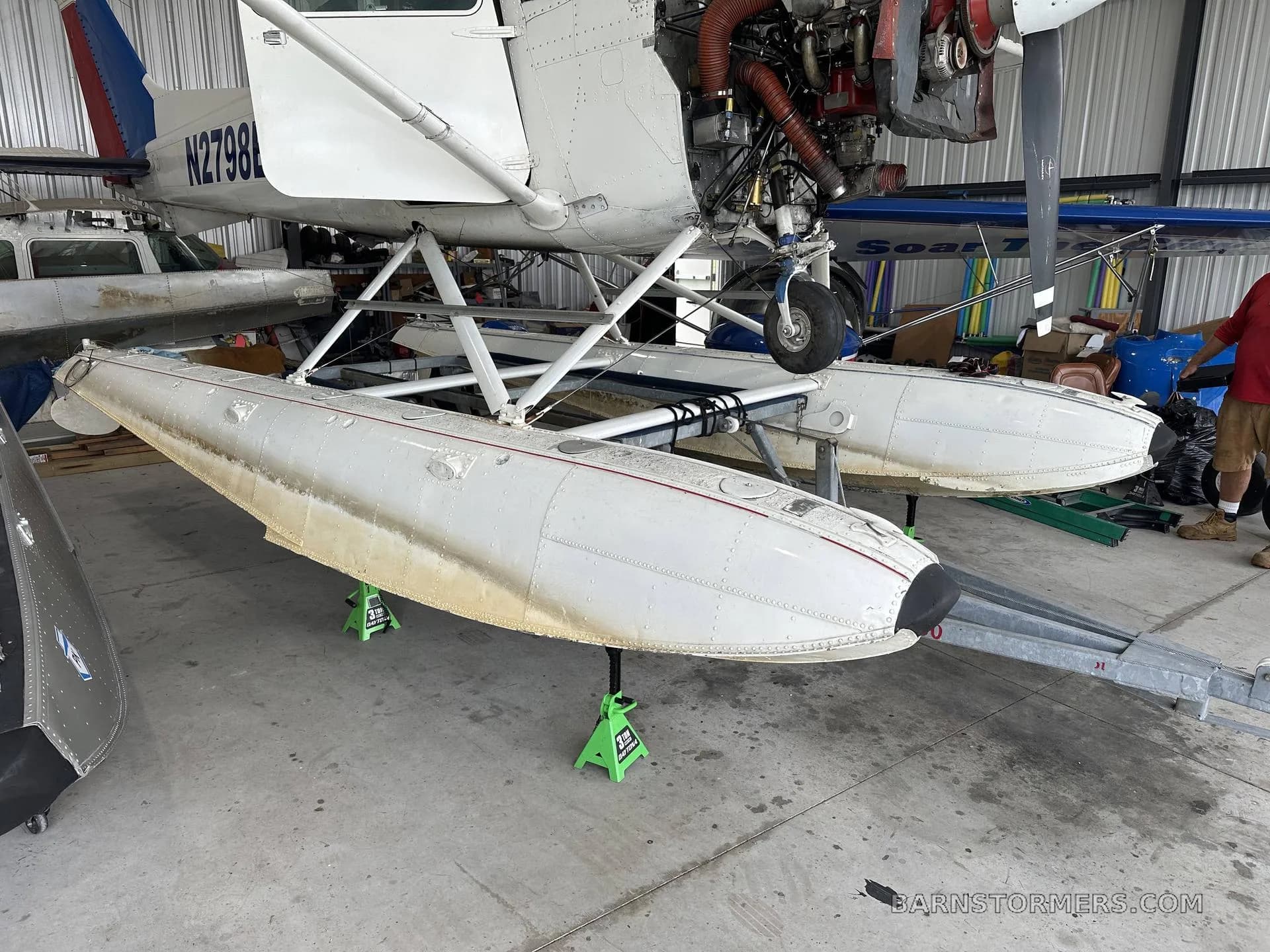 A white floatplane, registration N2798L, is seen on jacks in a hangar, with its engine cowling open.