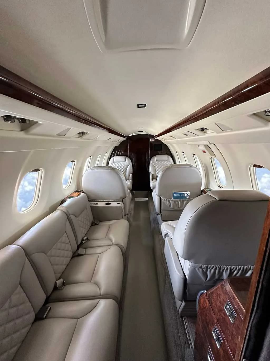 The luxurious interior cabin of a private jet, featuring light grey quilted leather seats and dark wood accents.
