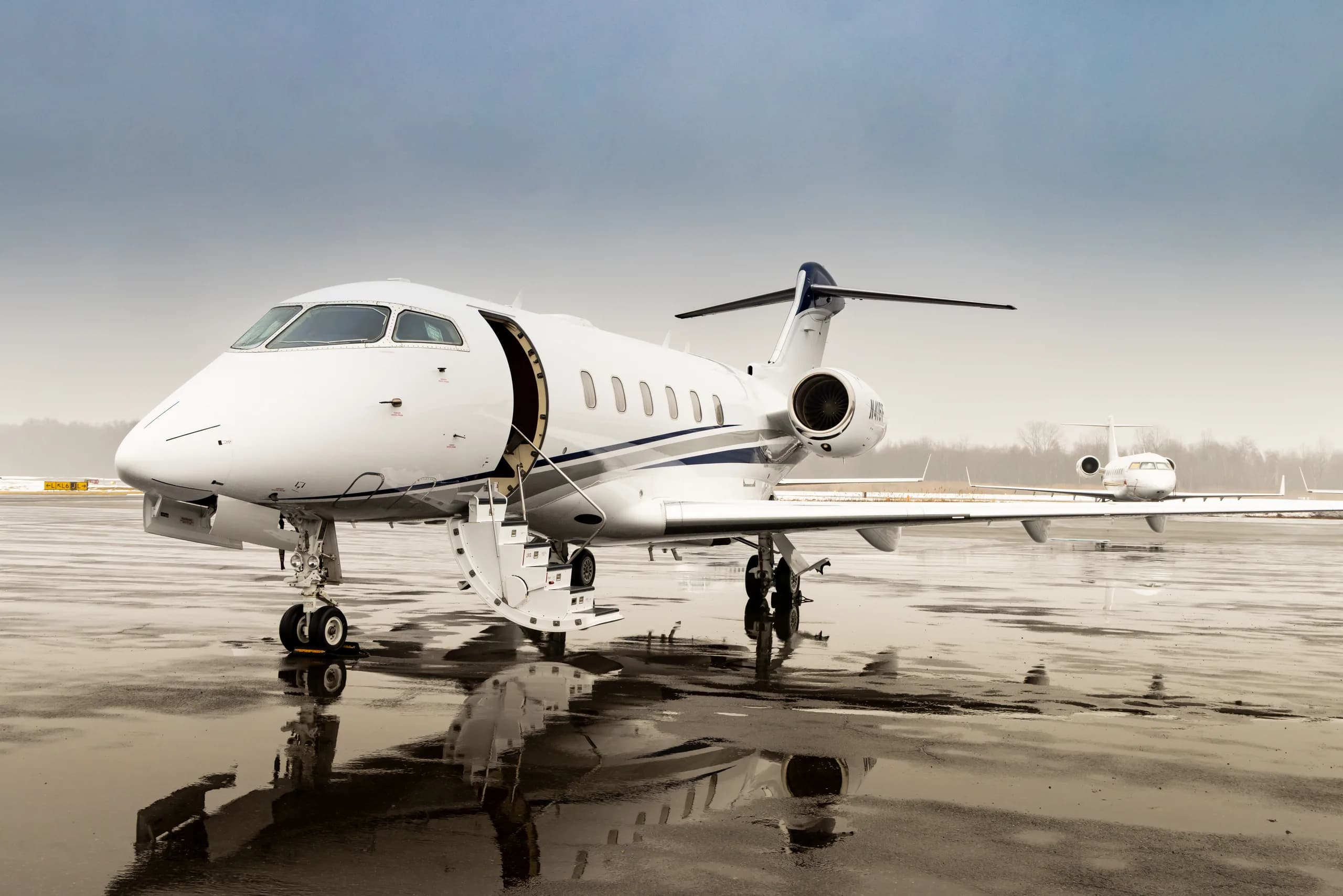 A white Bombardier Challenger 300 business jet with blue and grey stripes on a wet tarmac, with its main door open.