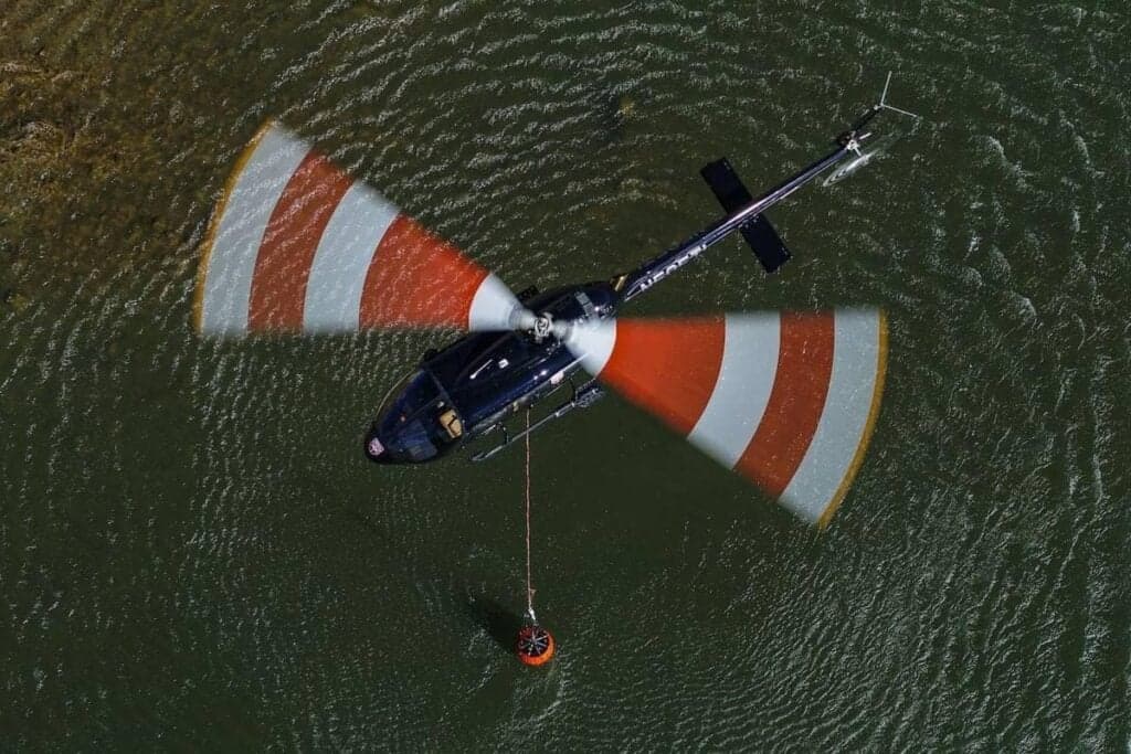 Overhead view of a black helicopter with red and white striped rotor blades carrying a water bucket over water.