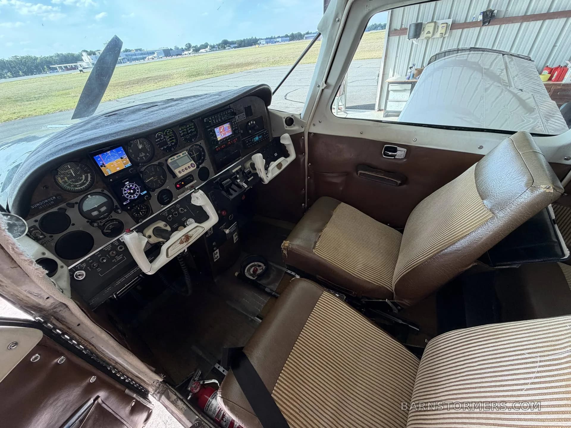 The interior of a small aircraft showing the cockpit and two brown and tan striped seats, with an airfield visible through the windshield.