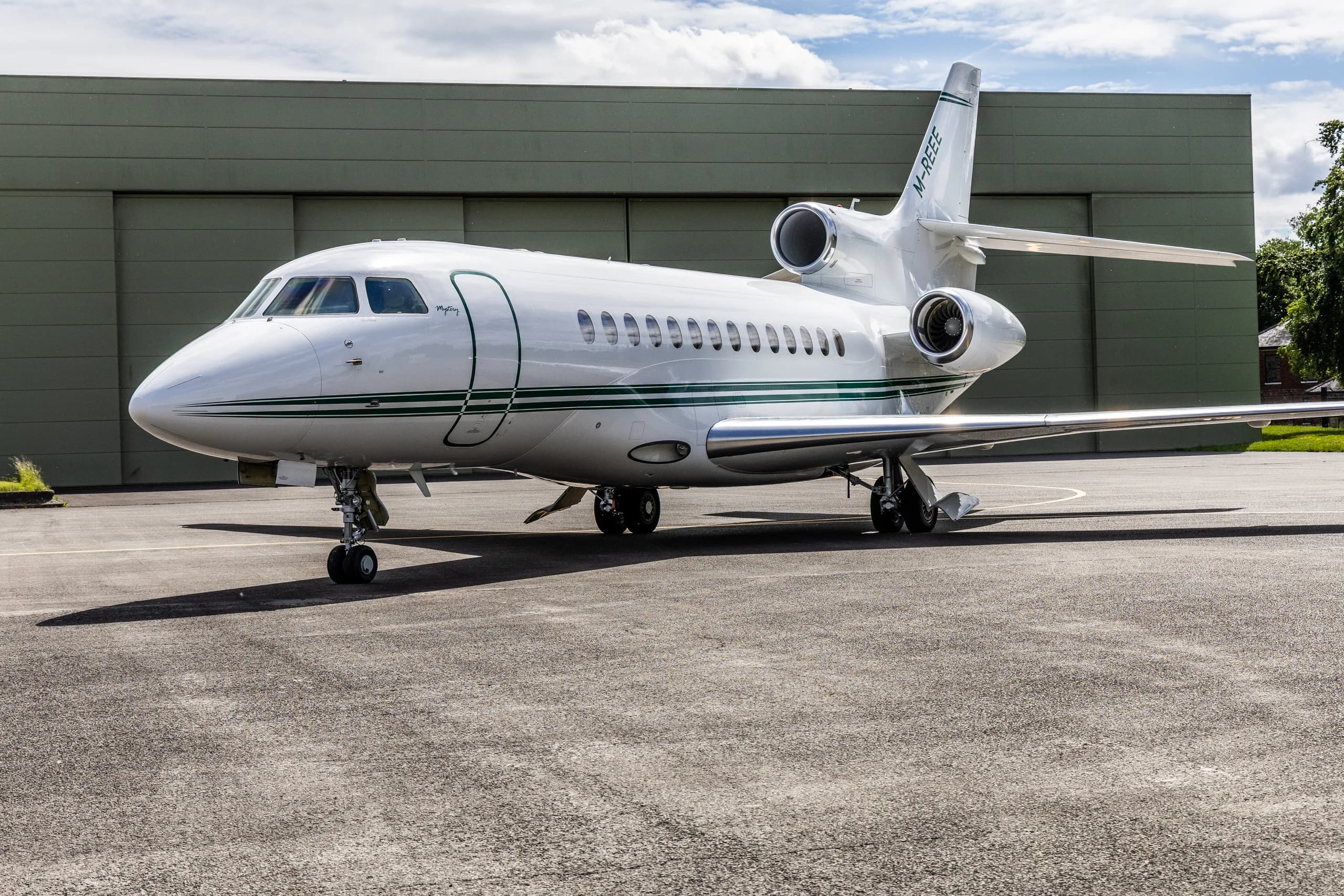 A white Dassault Falcon 900EX with a green stripe, registration M-REEE, parked on an airfield in front of a green hangar.