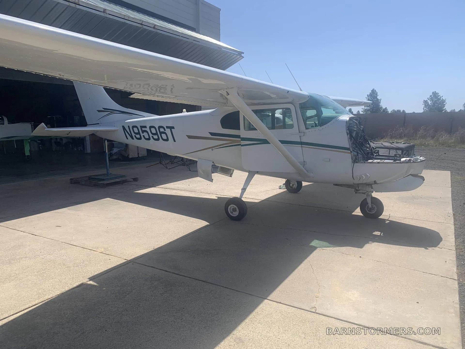 A white Cessna 182 aircraft with green and gold stripes, registration N9596T, parked on concrete outside a hangar with its engine cowling removed.