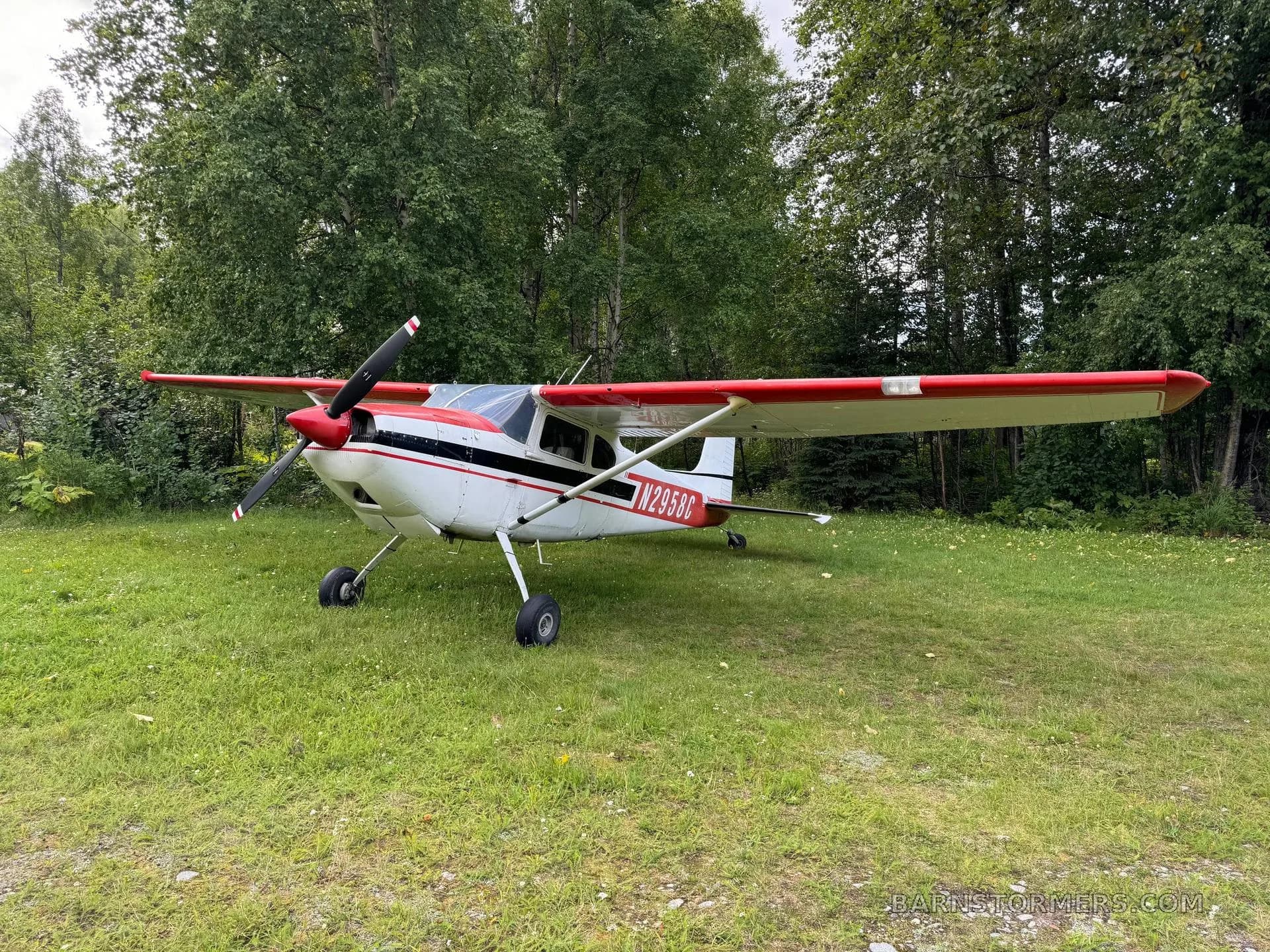 A white and red aircraft (N2958C) with conventional landing gear parked on a grassy field next to trees.