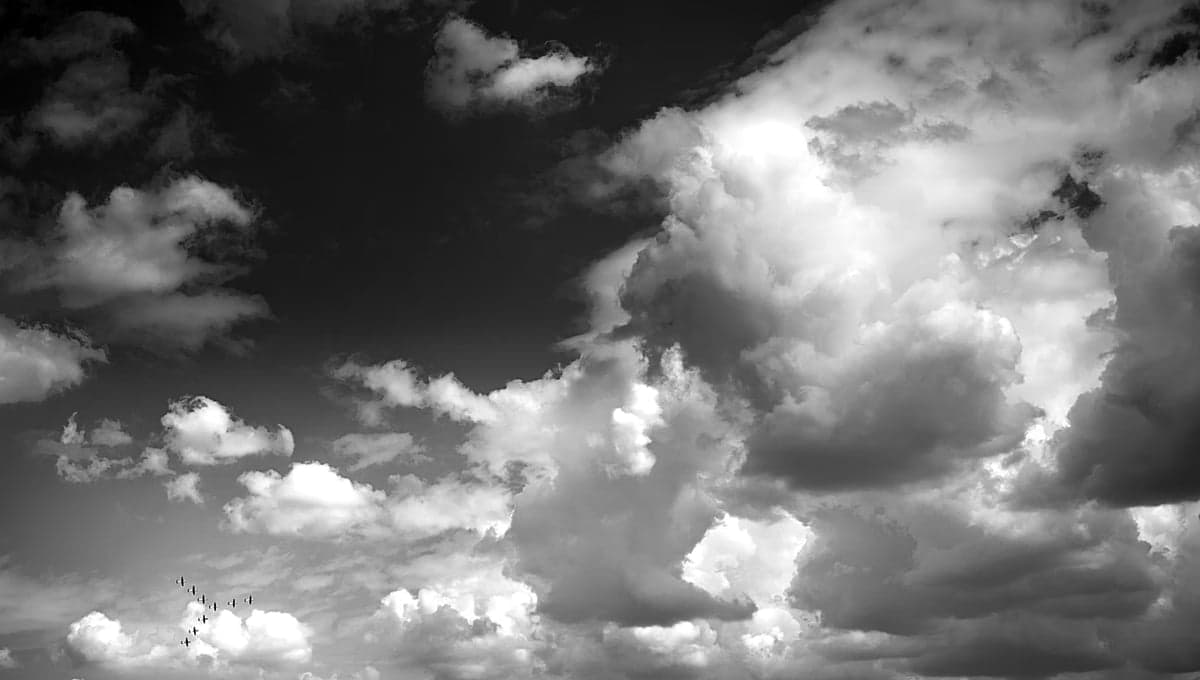 A black and white photo of a formation of small aircraft flying through a cloudy sky.
