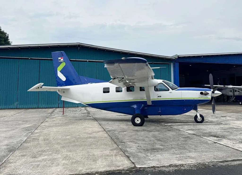 A blue and white Quest Kodiak 100 aircraft with a green stripe and Panamanian flag on the tail, parked on a concrete apron next to a hangar.