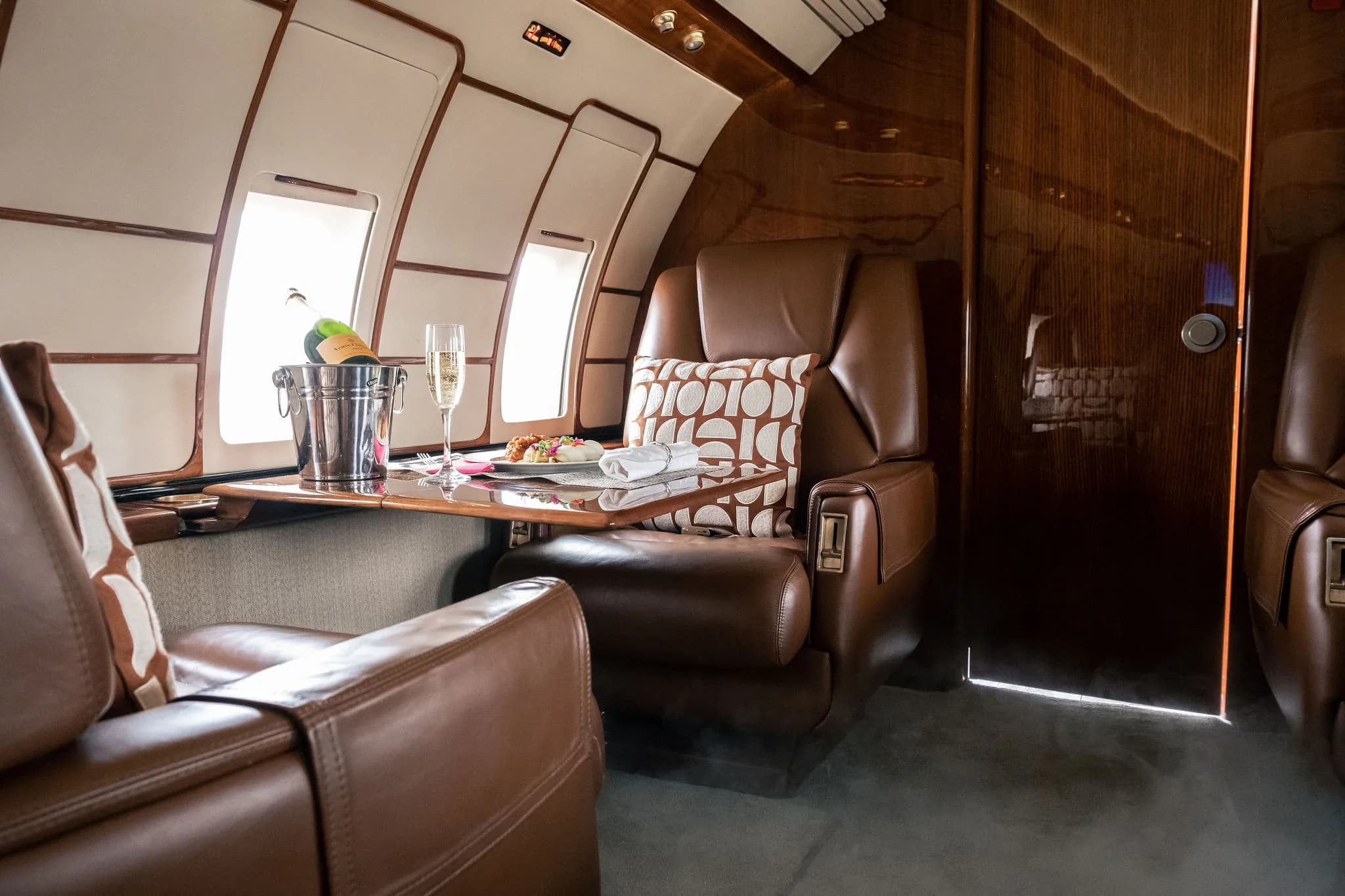 A private jet interior with brown leather seats and a table holding a champagne bucket and glass.