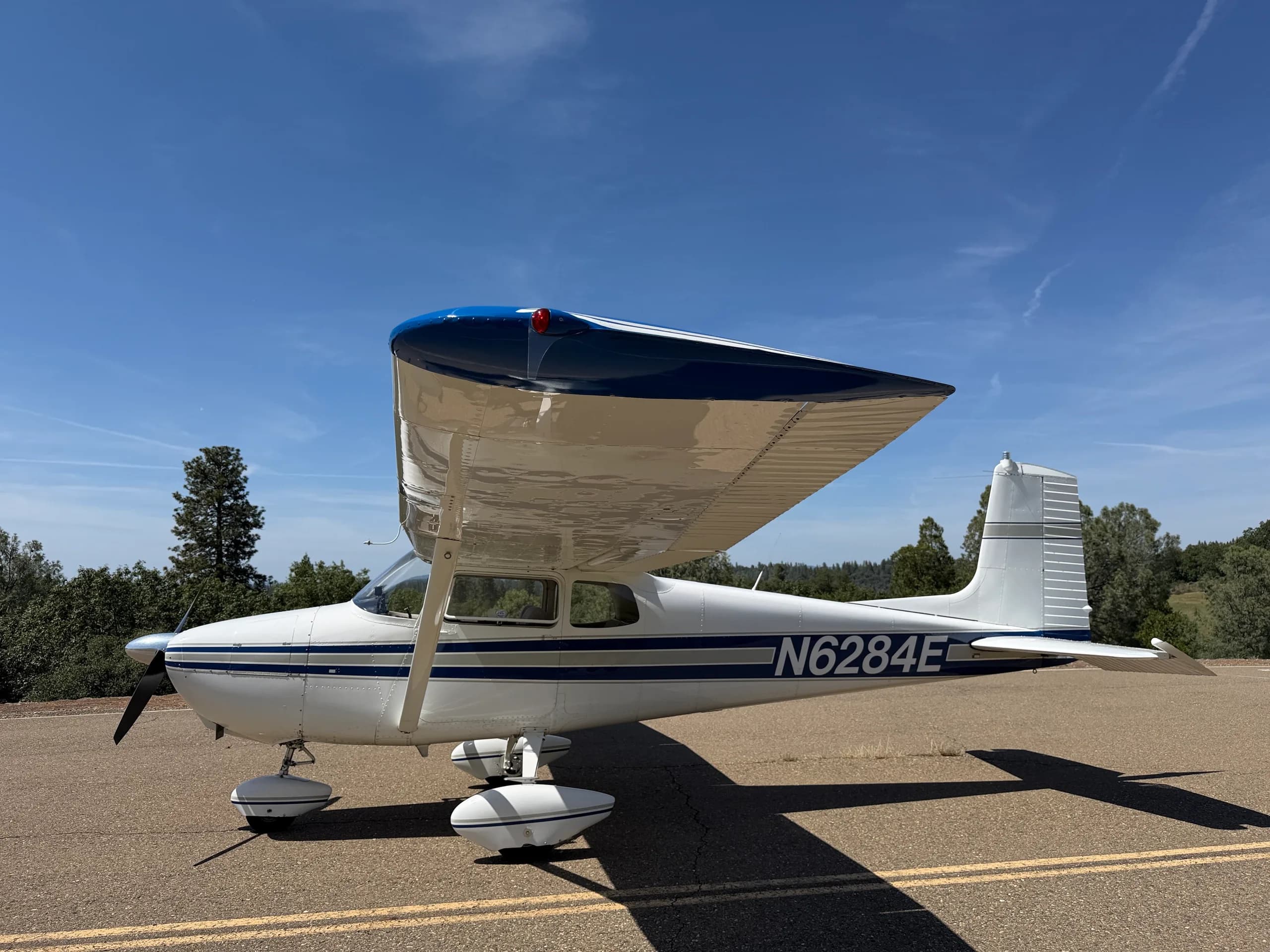 A side view of a white Cessna 172 with blue stripes, N6284E, parked on an asphalt surface with trees in the background.