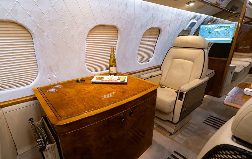 A close-up of a private jet's interior showing a wooden table with wine and snacks next to a beige seat.
