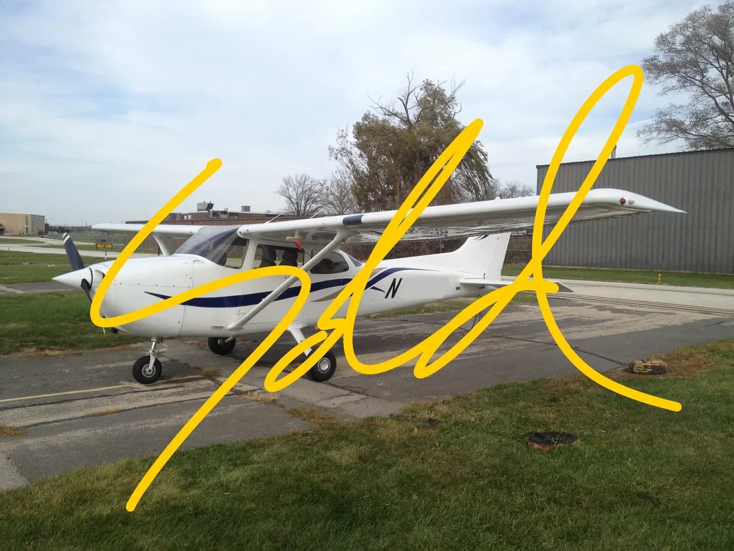 A white Cessna 172-like aircraft with blue stripes is parked on an asphalt ramp, with a building in the background.