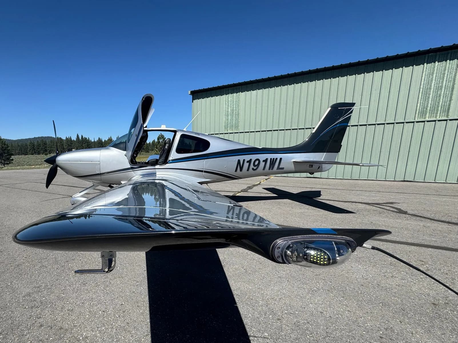 A low-angle view of the right wing and fuselage of a silver and black Cirrus SR22T aircraft with N191WL, with the door open.