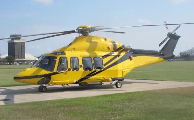 A yellow AgustaWestland AW139 helicopter with black stripes parked on a tarmac with a control tower in the background.