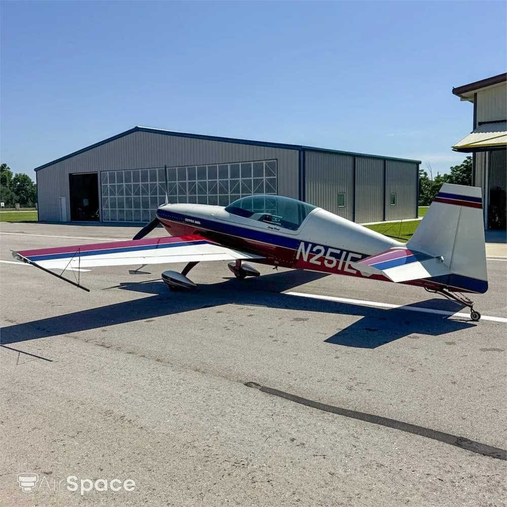 A side view of the red, white, and blue Extra 300L aircraft, N251EC, parked on an airfield with a hangar in the background.
