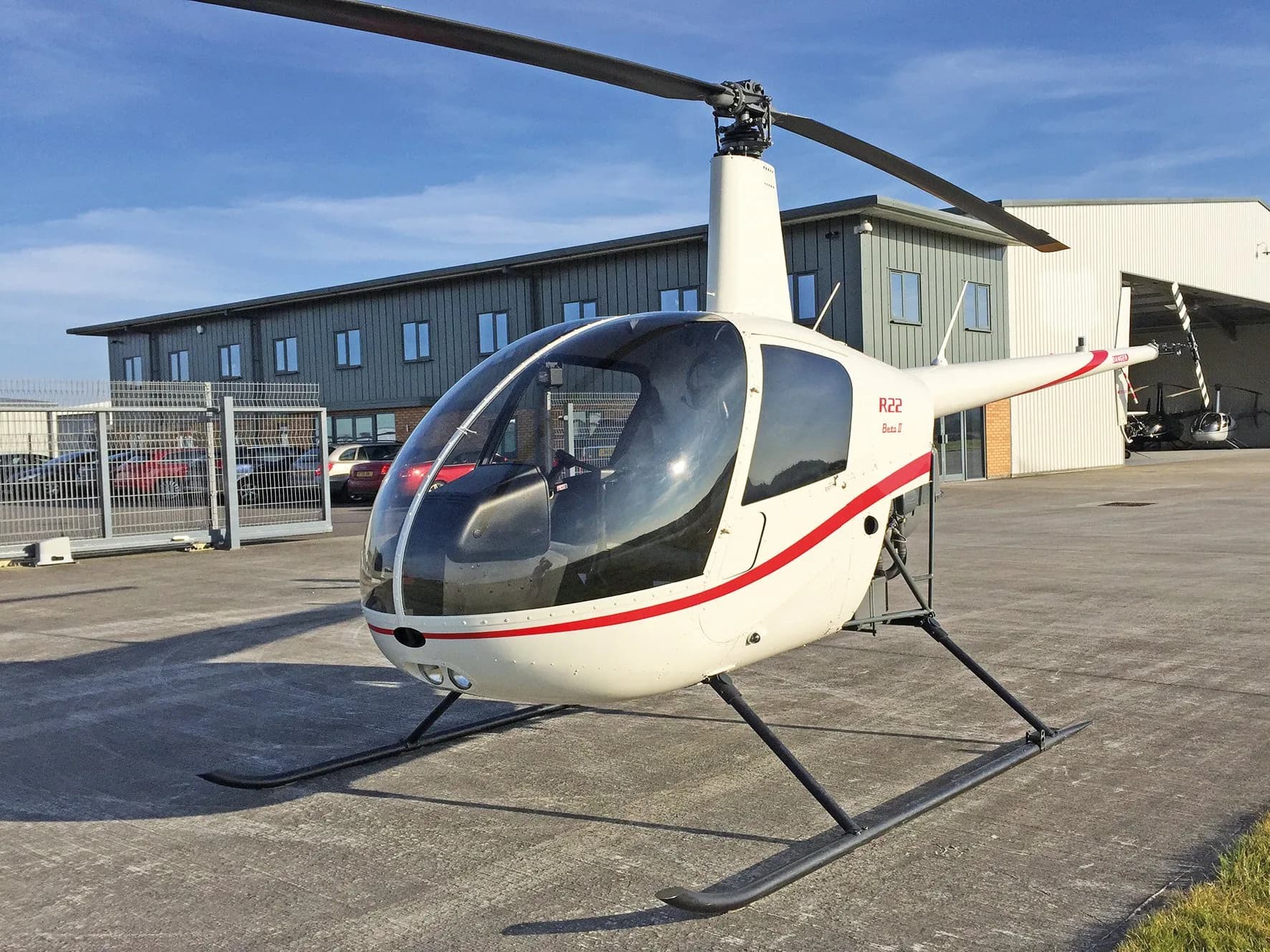 A white Robinson R22 Beta II helicopter with red stripes is parked on a tarmac in front of a modern building.