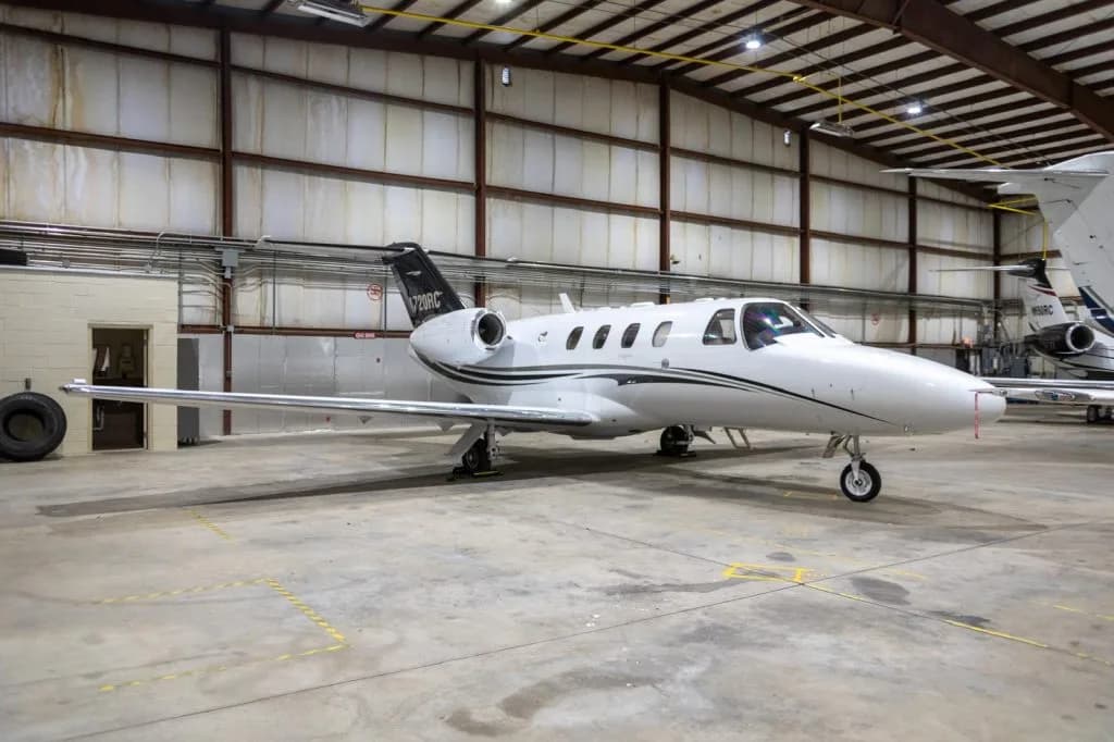 A white Cessna Citation M2 business jet with black and grey stripes is parked inside a hangar.