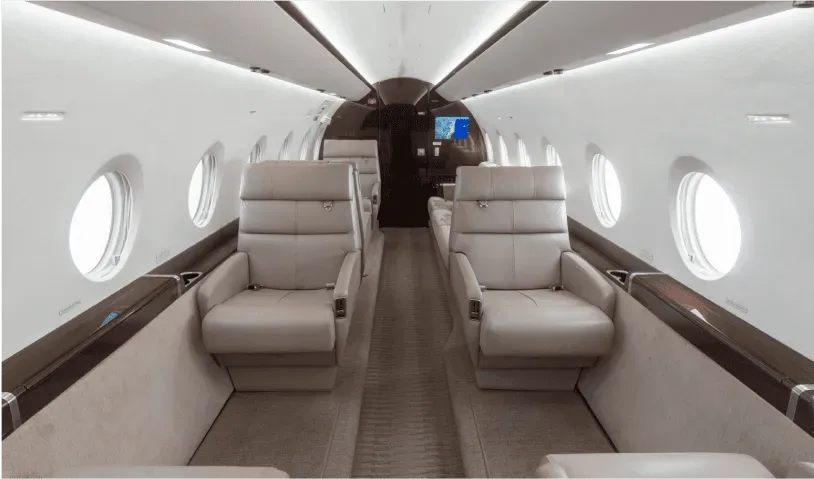 View down the aisle of a luxurious aircraft cabin with light grey leather seats and large oval windows.