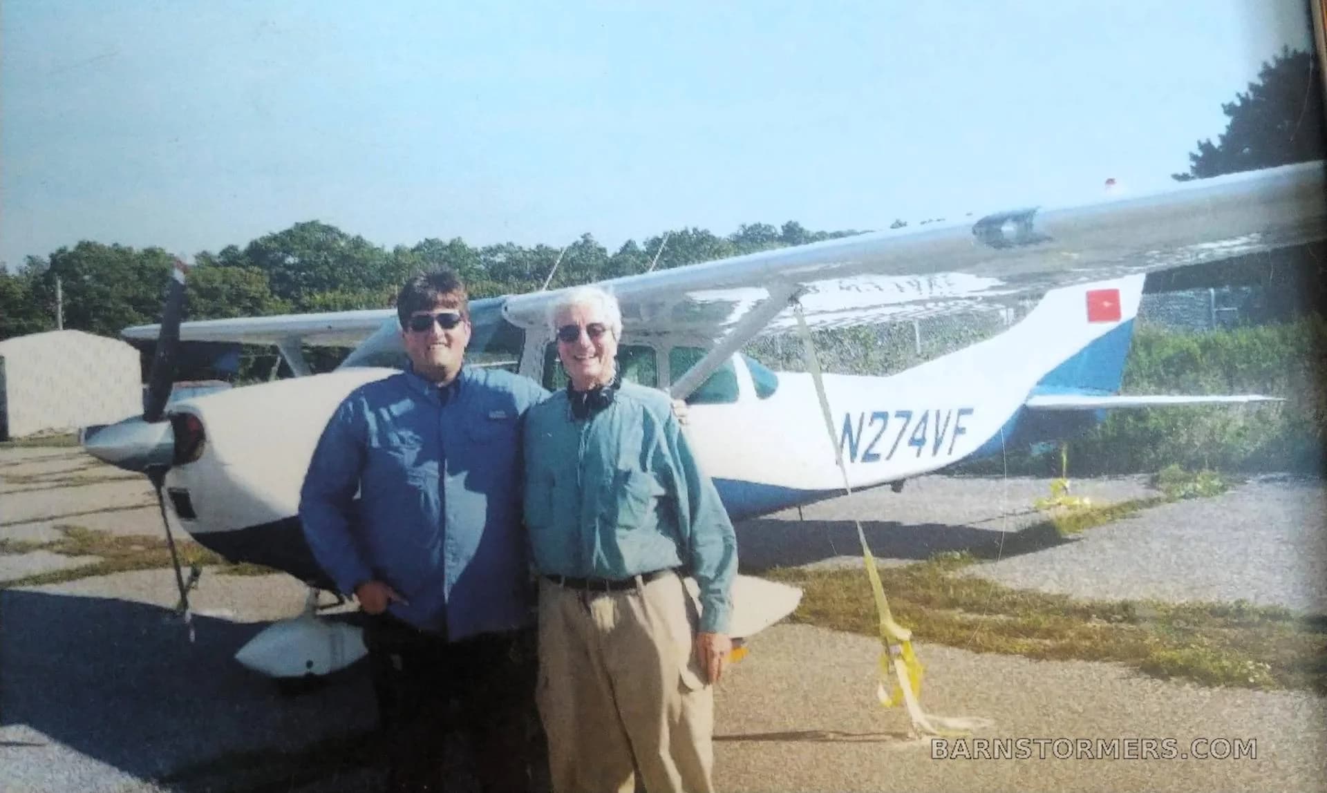 Two men standing in front of a white and blue single-engine aircraft (N274VF) parked on an asphalt ramp.