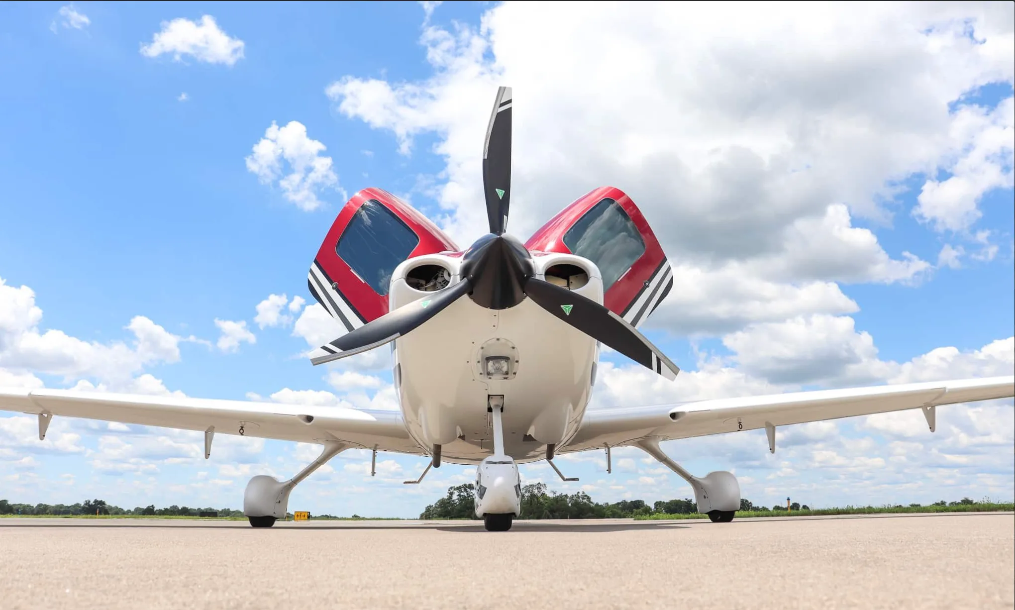 A low-angle front view of a white and red single-engine aircraft with open gull-wing doors on an airfield.