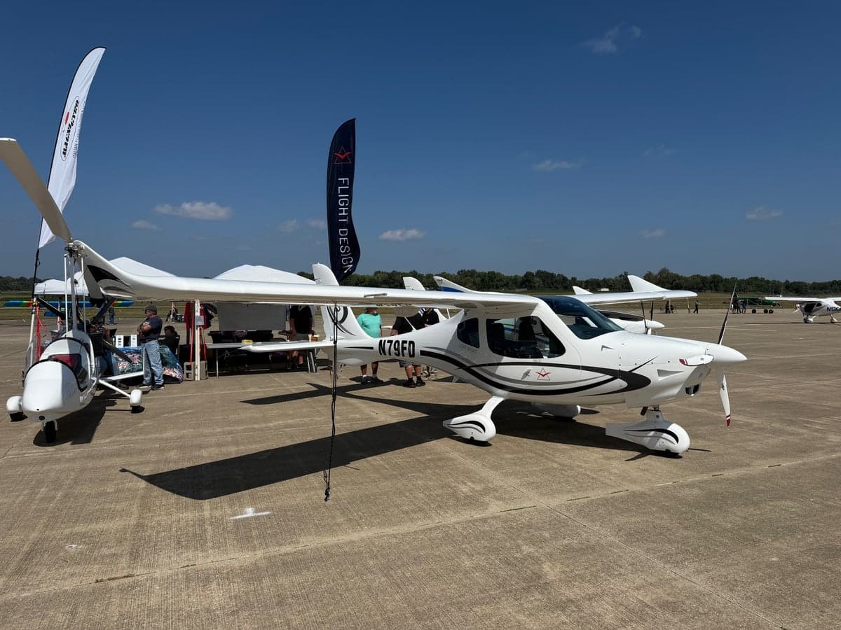 A white Flight Design light sport aircraft (N79FD) with black and grey decals parked on an airfield tarmac, with an autogyro and vendor tents nearby.