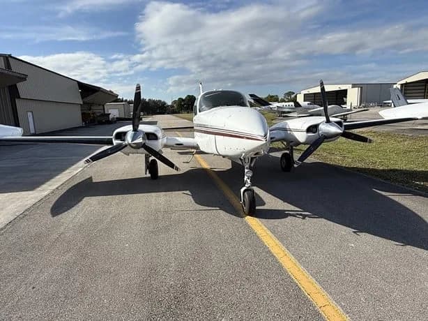 Front view of a white twin-engine aircraft with red and black stripes, parked on an airfield.