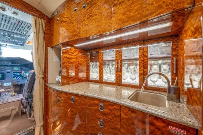 A luxurious aircraft galley with polished wood cabinetry, a granite countertop, a sink, and glassware storage, with a view into the cockpit.