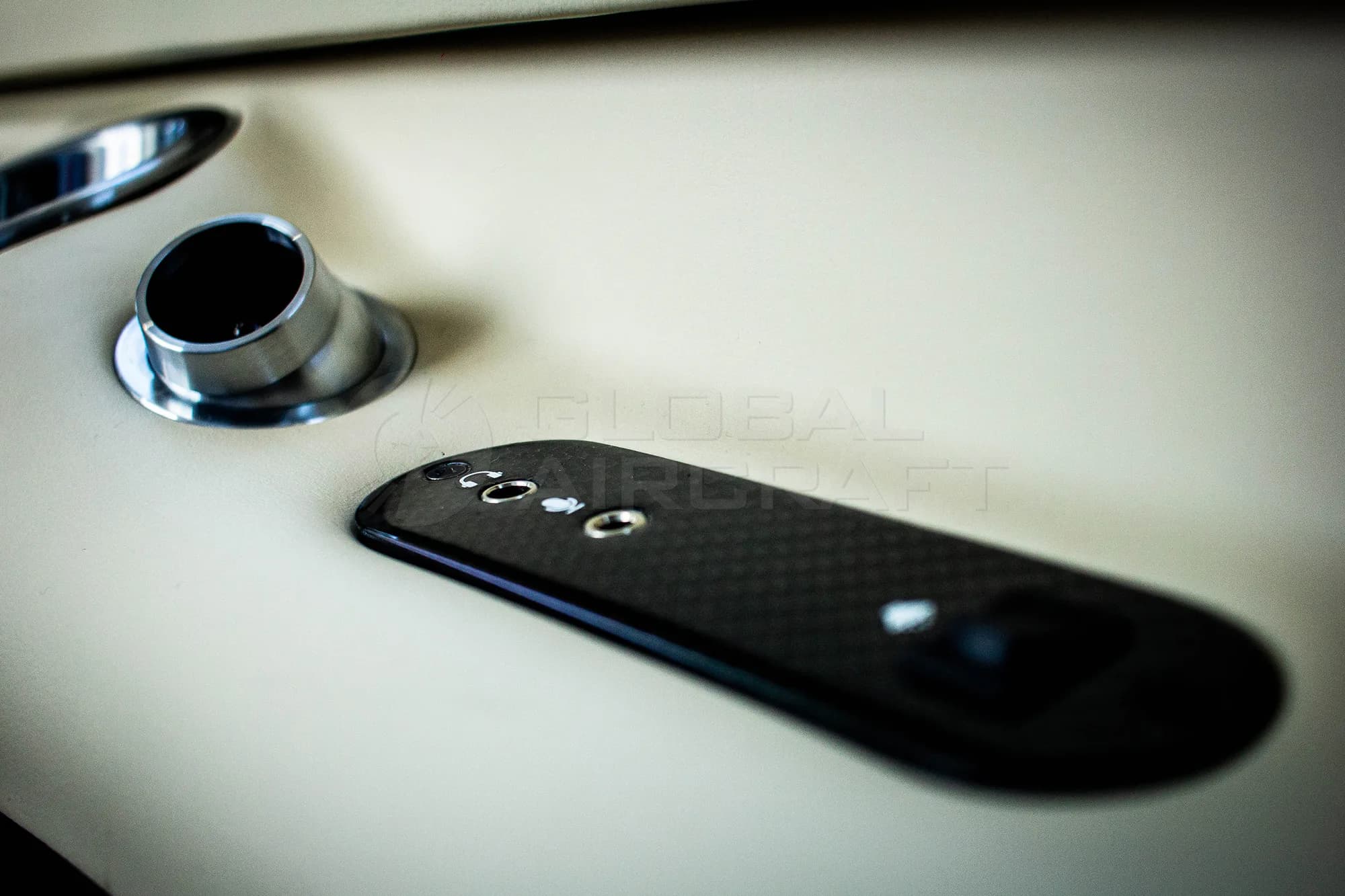A close-up of an aircraft interior detail, showing a chrome cup holder and a black carbon fiber panel with buttons.