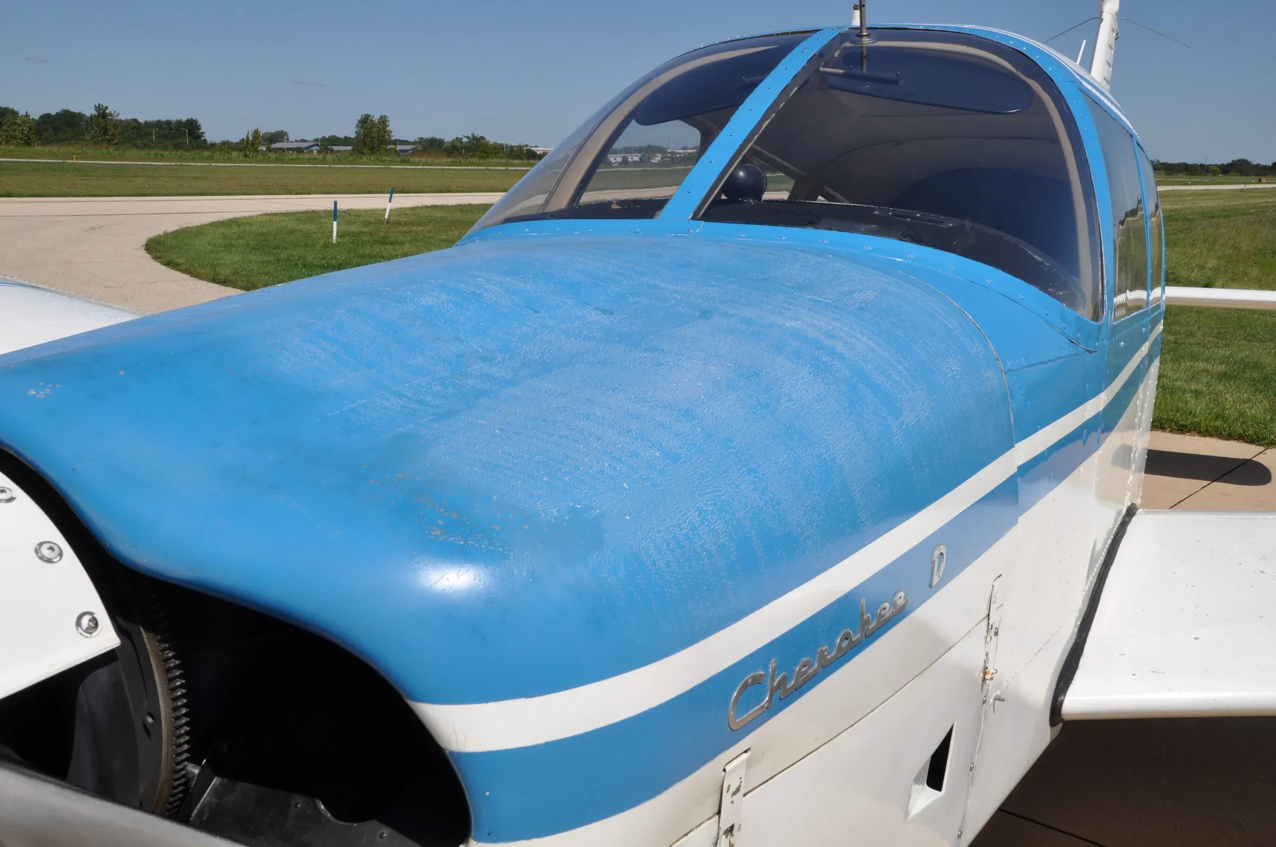 A close-up view of the blue and white nose and cockpit of a Piper Cherokee D aircraft, showing the 'Cherokee D' emblem.