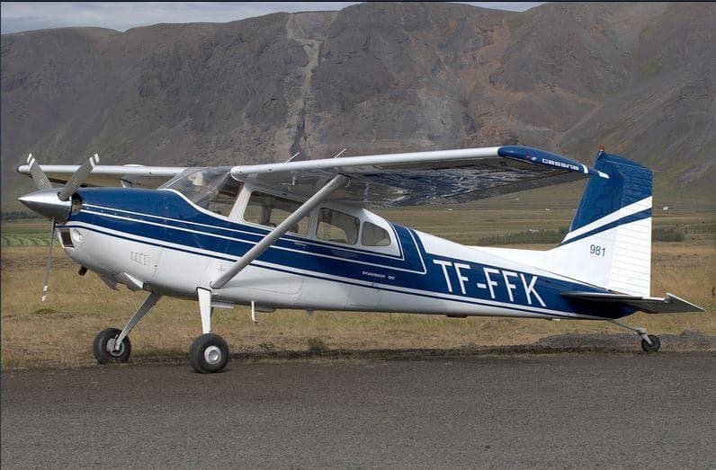 A white and blue Cessna 180 aircraft, registration TF-FFK, parked on an airfield with mountains in the background.