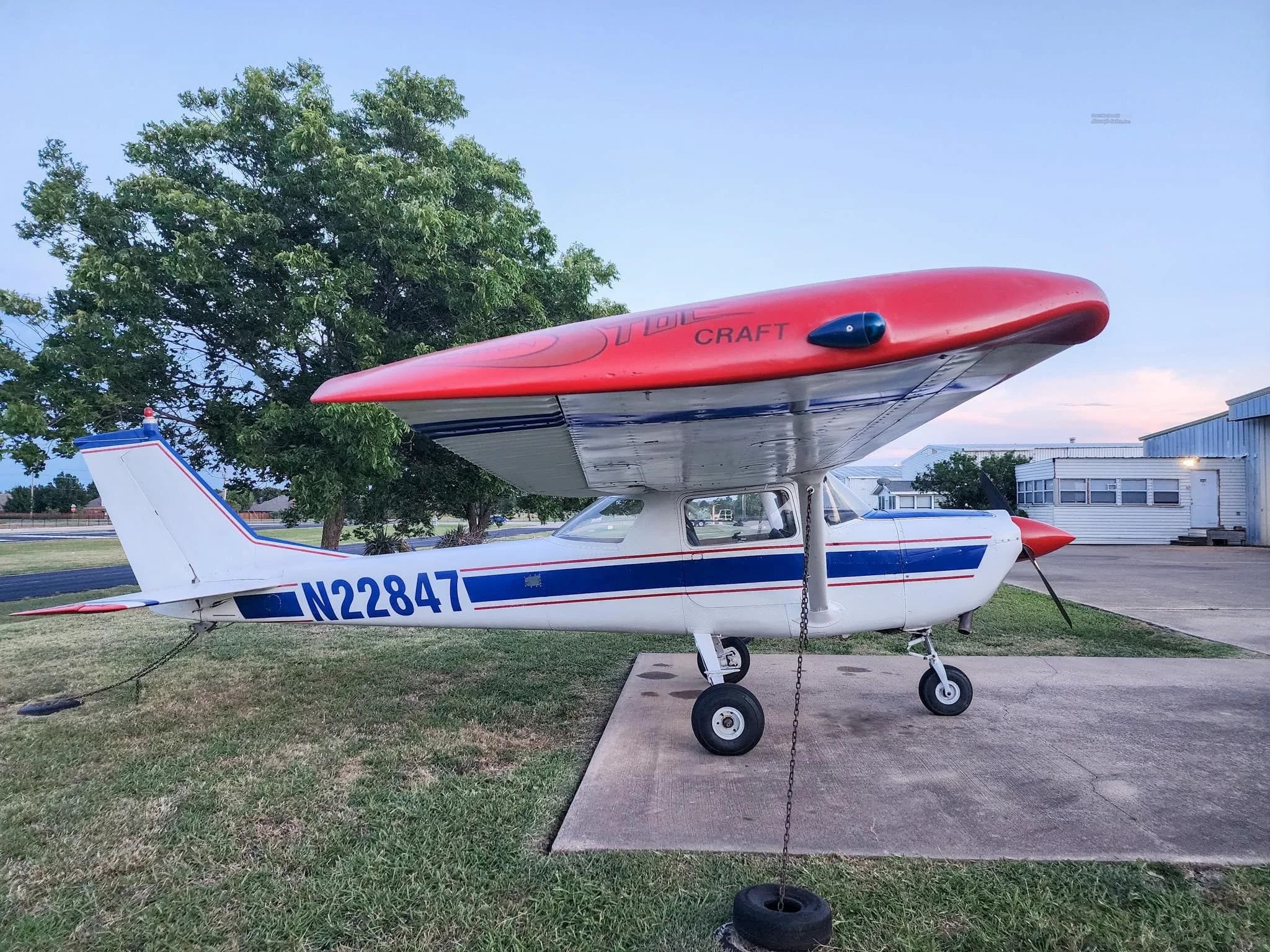 A white and blue Cessna 172 with a red wing fairing is parked on a concrete pad with a hangar in the background.