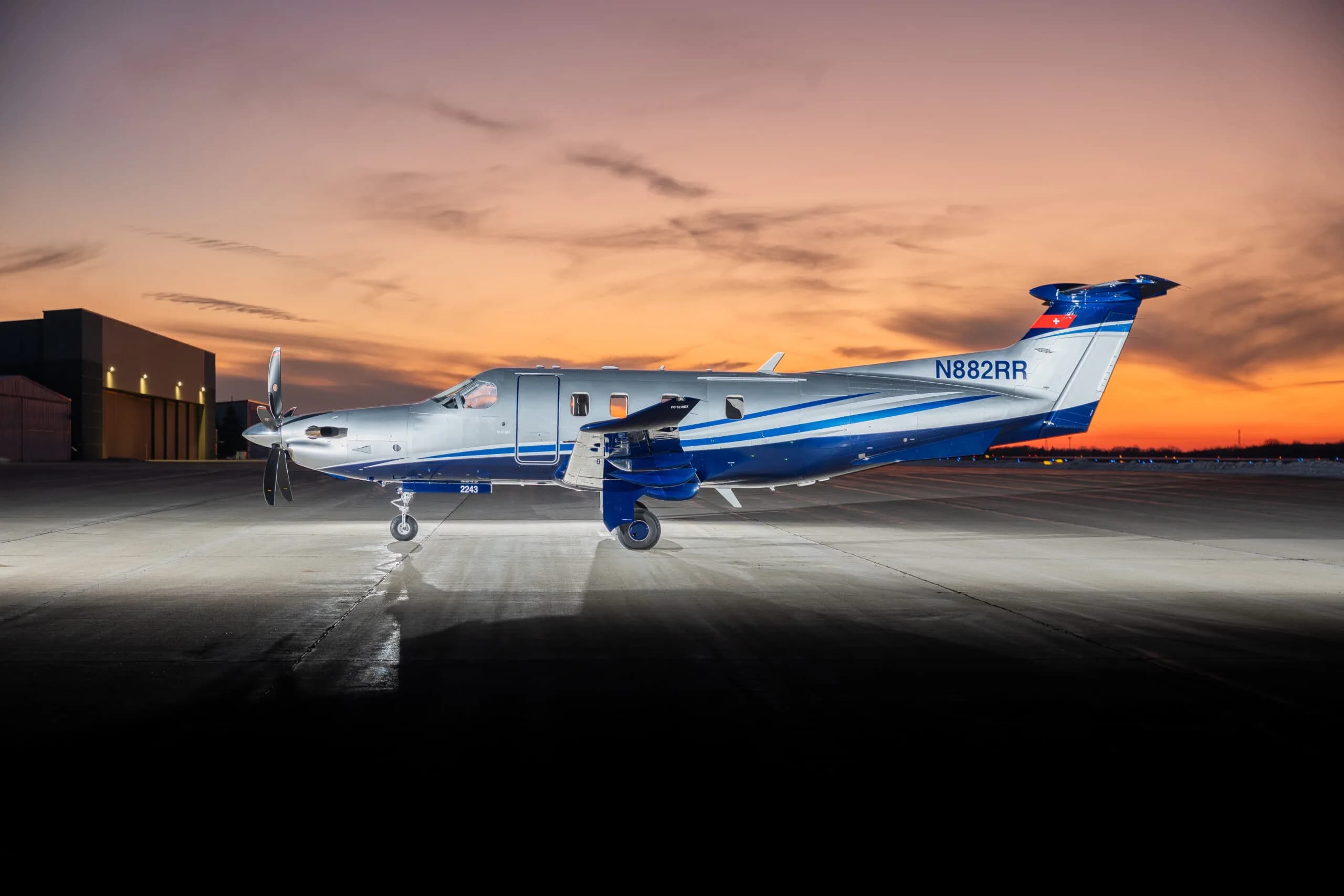 A silver and blue Pilatus PC-12 turboprop aircraft with registration N882RR parked on a tarmac with a hangar in the background at dusk.