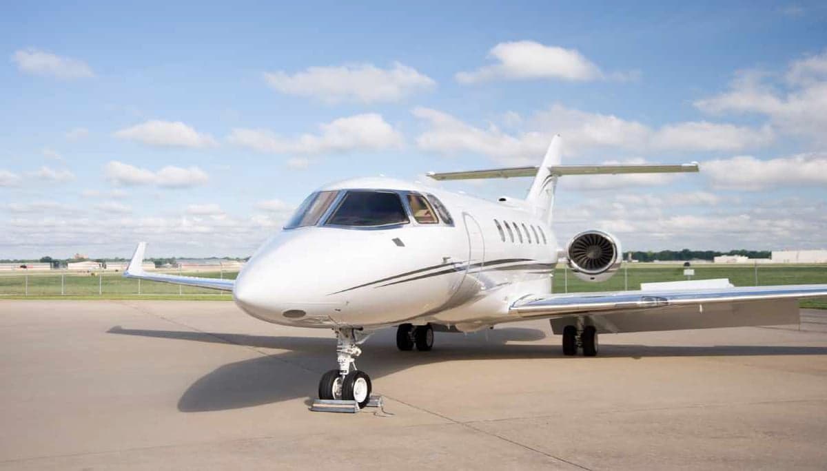 A white business jet with black and grey stripes parked on an airfield under a blue sky.