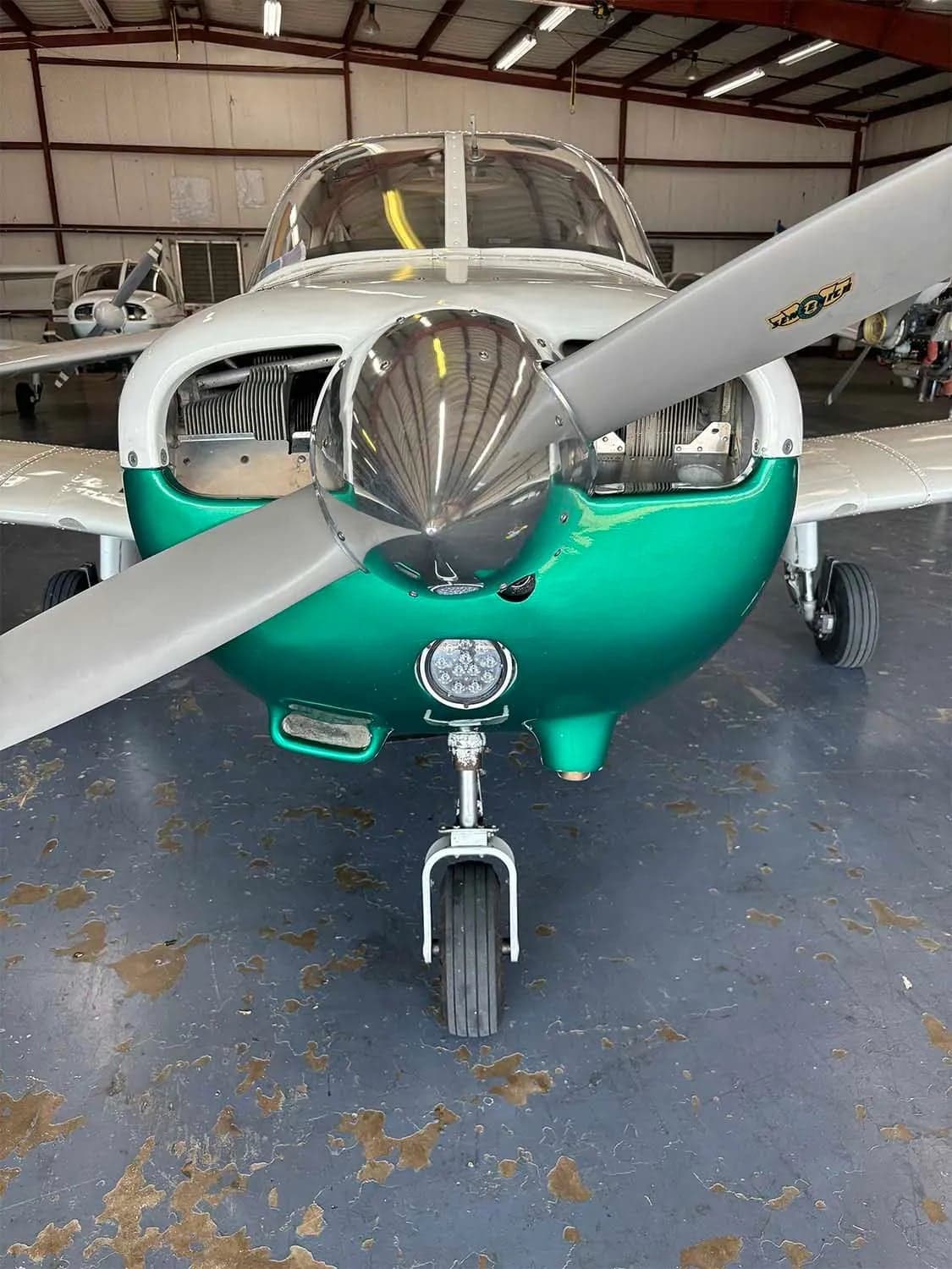 A front view of a green and white single-engine aircraft with its engine cowling open in a hangar.