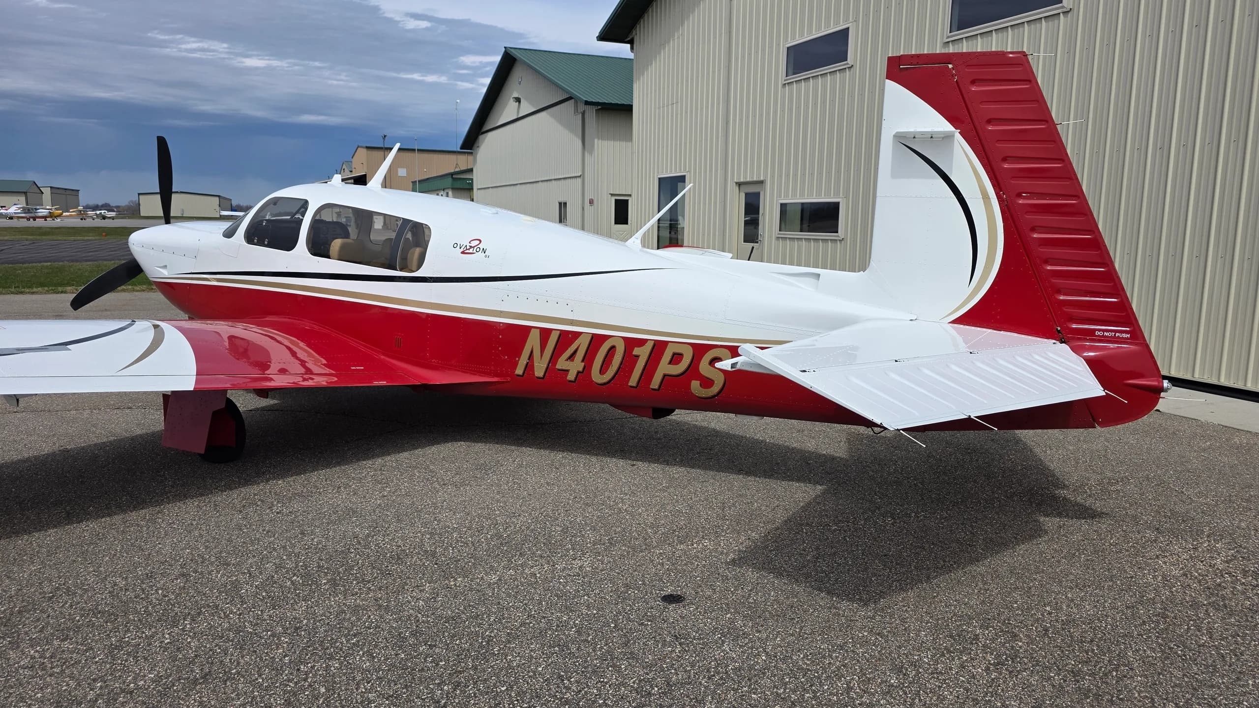 A side view of a red and white Mooney aircraft with registration N401PS parked on an asphalt apron next to a hangar.