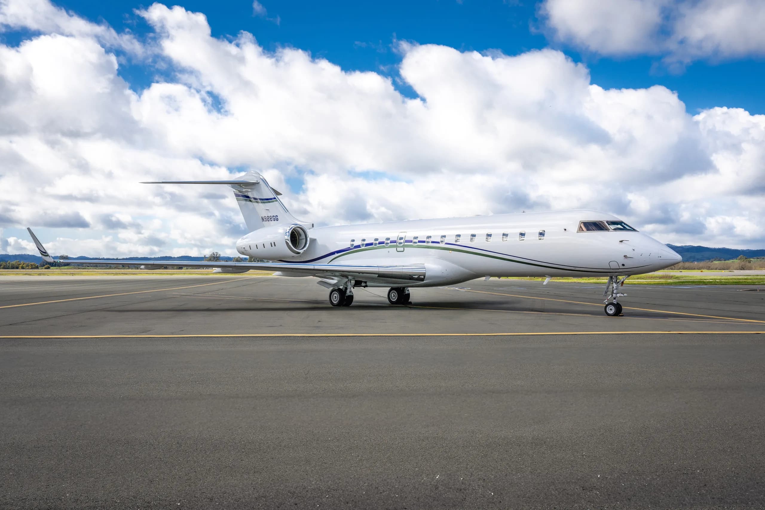 A white Bombardier Global 6000 business jet with blue and green stripes, parked on an airport tarmac with hills in the background under a blue sky.