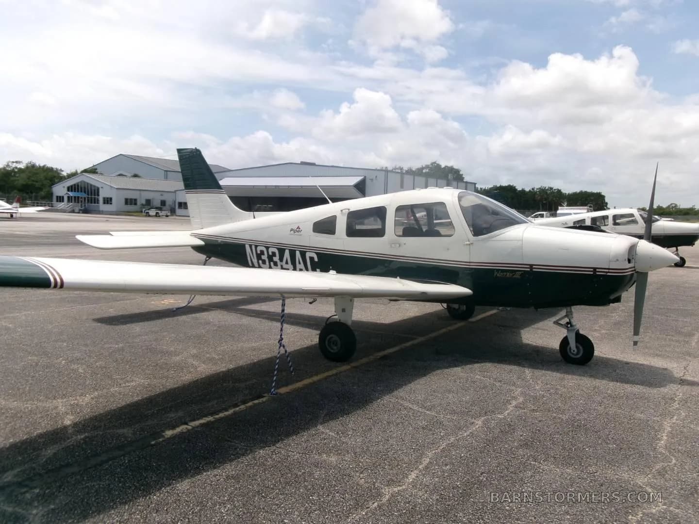 A side view of a white and dark green Piper Archer II (N334AC) parked on an airfield with hangars in the background.