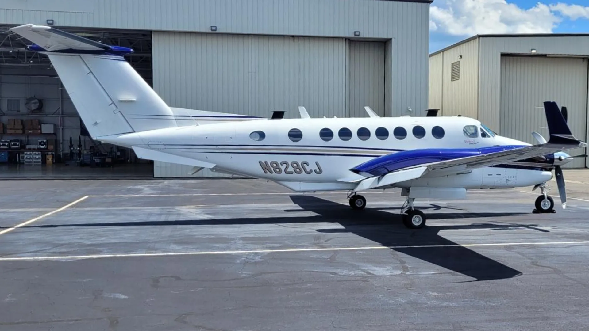 A white and blue Beechcraft King Air N828CJ parked on an asphalt tarmac with hangars in the background.