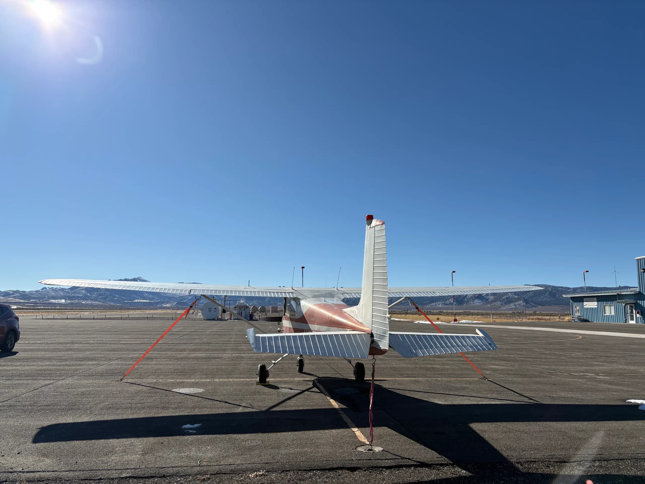 A red and white single-engine high-wing aircraft, N5799A, viewed from directly behind, parked on an airfield under a bright sun.