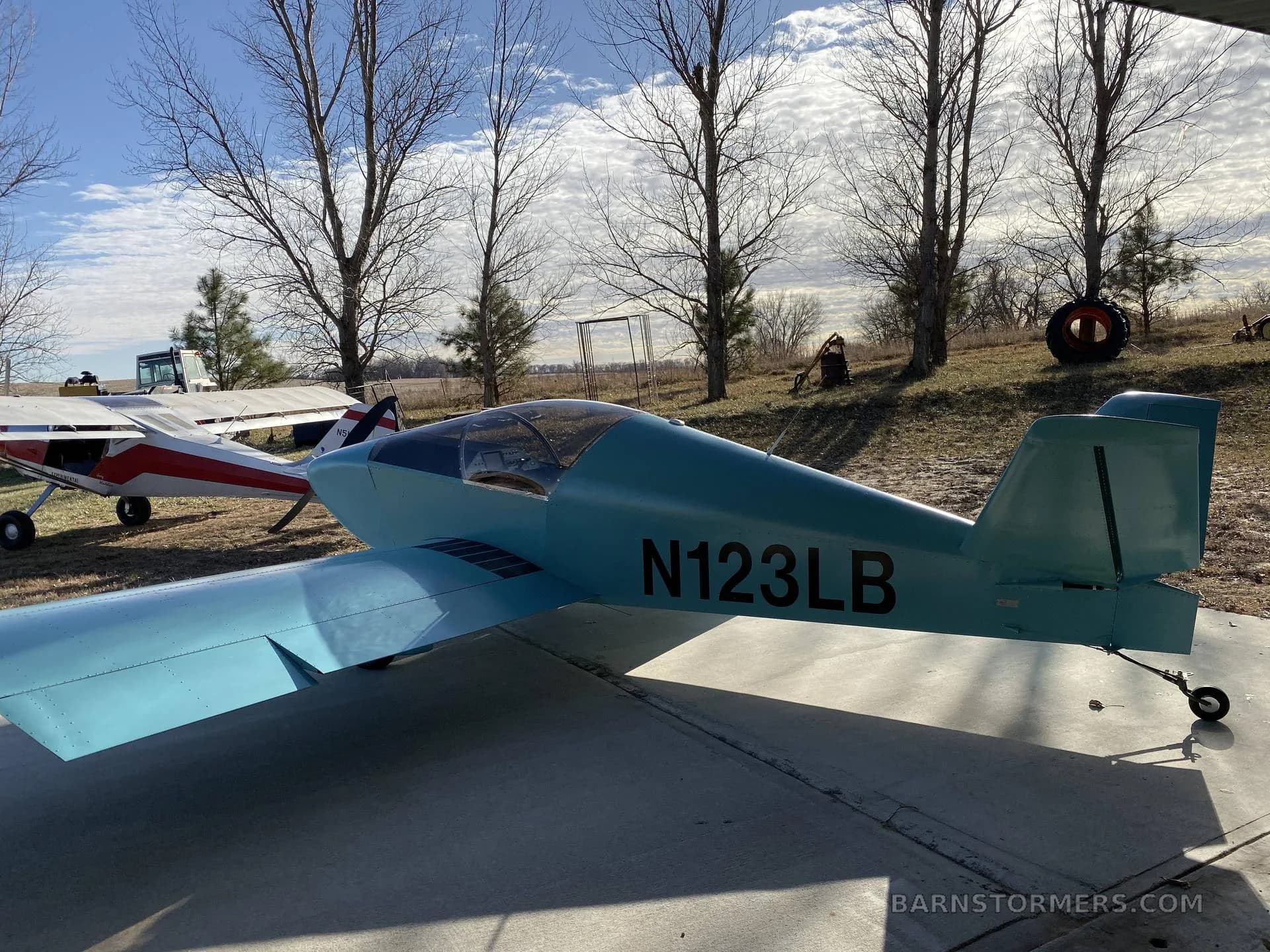 A light blue single-engine experimental aircraft (N123LB) parked on concrete with another white aircraft behind it and bare trees in the background.