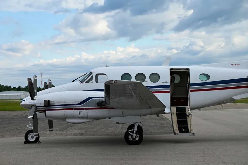 A side view of a white Beechcraft King Air C90GTx with blue and red stripes, parked on the tarmac with its passenger door open.