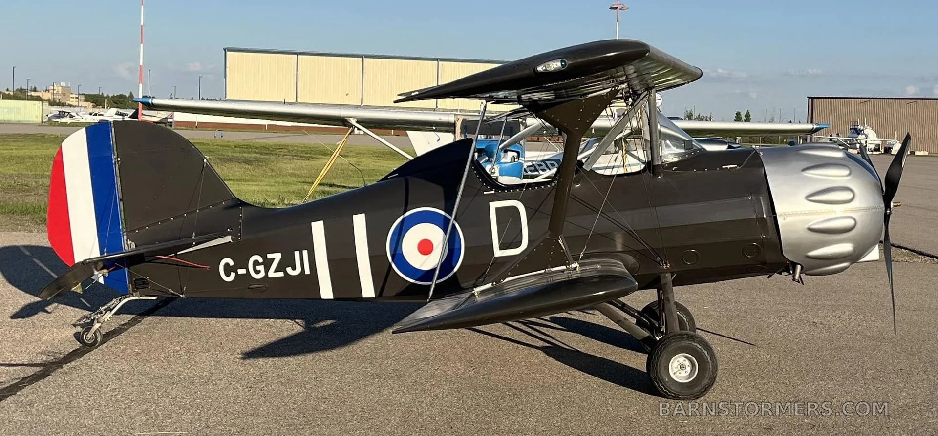 A black biplane with a silver engine cowling and British roundel markings, parked on an airport tarmac with hangars in the background.