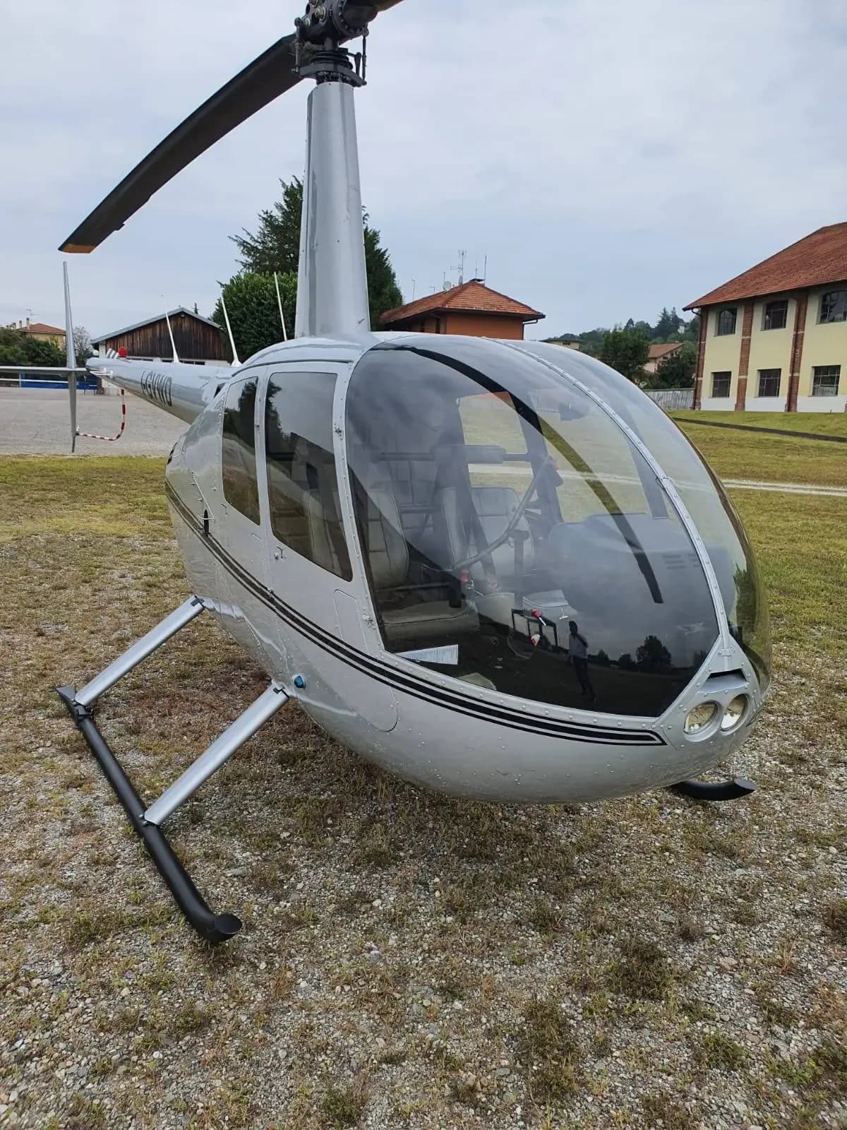 A silver Robinson R44 helicopter parked on a grassy field with buildings in the background.