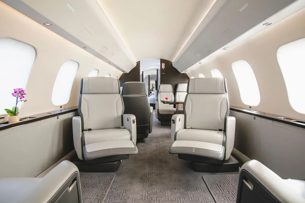 The spacious interior cabin of a private jet, showing rows of light grey and black leather seats and a patterned carpet.