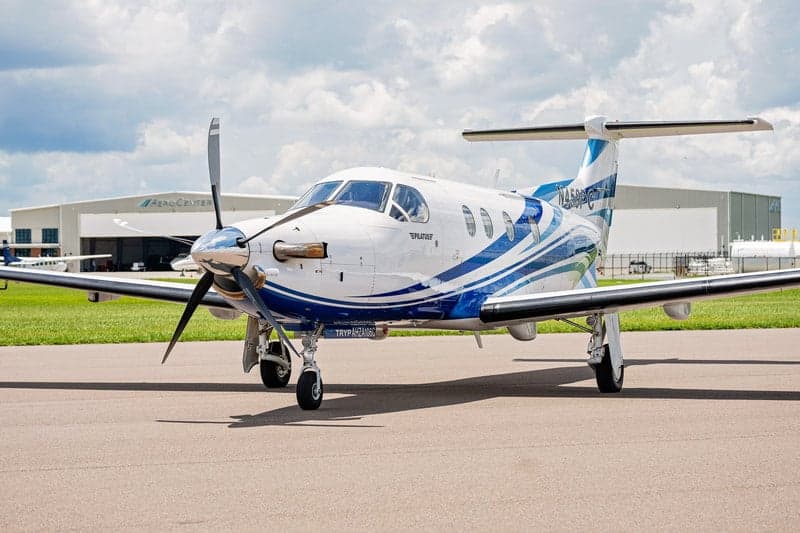 A Pilatus PC-12 aircraft with a blue and white paint scheme is parked on the tarmac in front of a hangar.