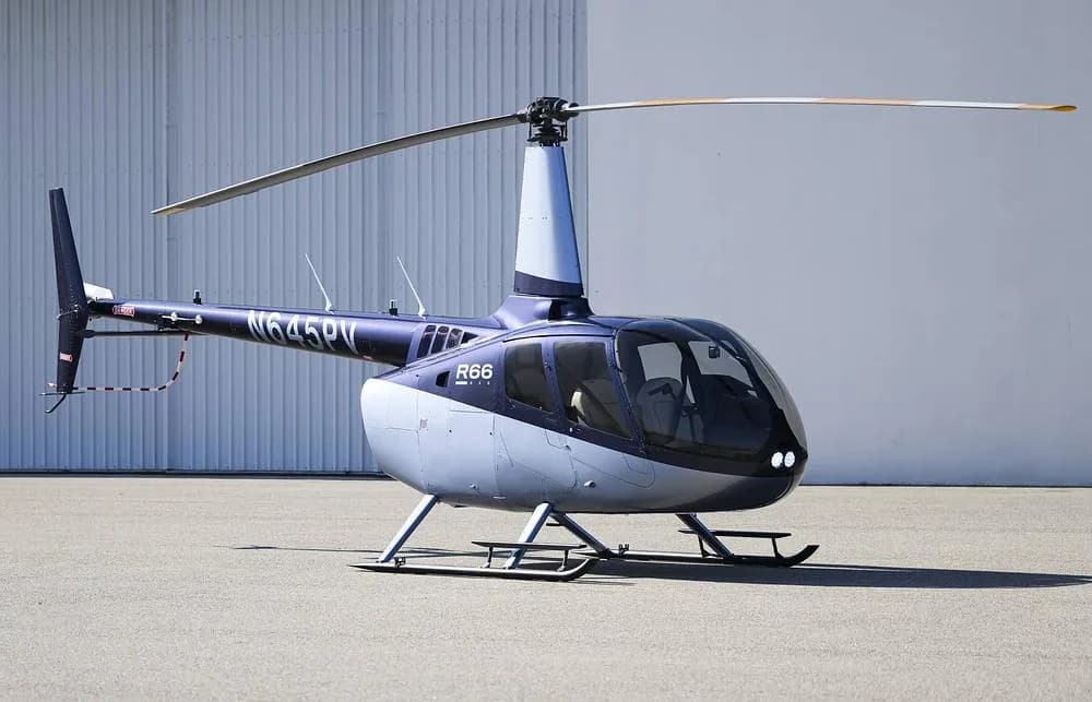 A blue and silver Robinson R66 helicopter (N645PV) is parked on an asphalt surface next to a white hangar.