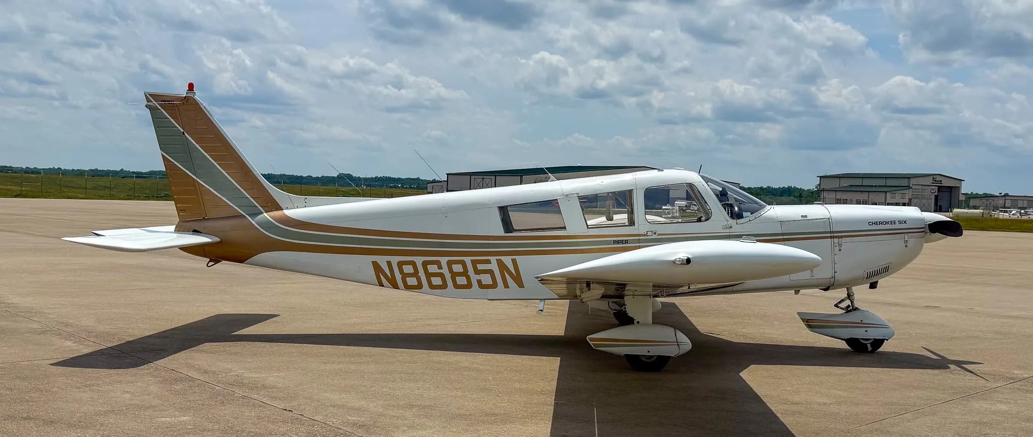 A side-right view of a white Piper Cherokee Six aircraft with brown and green stripes, showing registration N8685N, parked on an airfield.