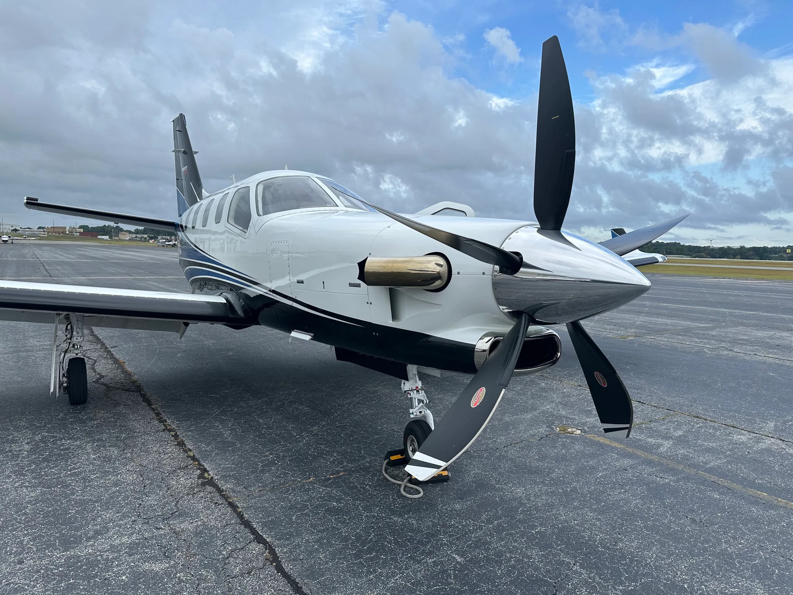 A white and blue TBM 960 turboprop aircraft with its propeller, parked on an asphalt tarmac under a cloudy sky.